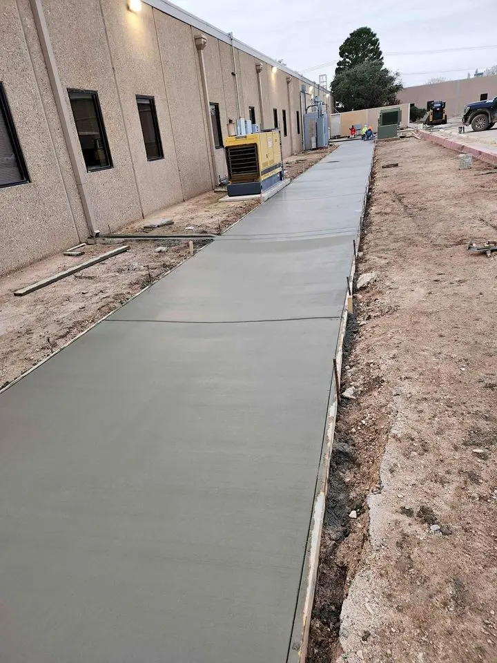 A freshly paved concrete walkway crafted by Bucks Concrete Craft runs alongside a building, bordered by dirt and construction materials. An industrial generator is visible on the left, and the overcast scene captures a day in Buckeye AZ.