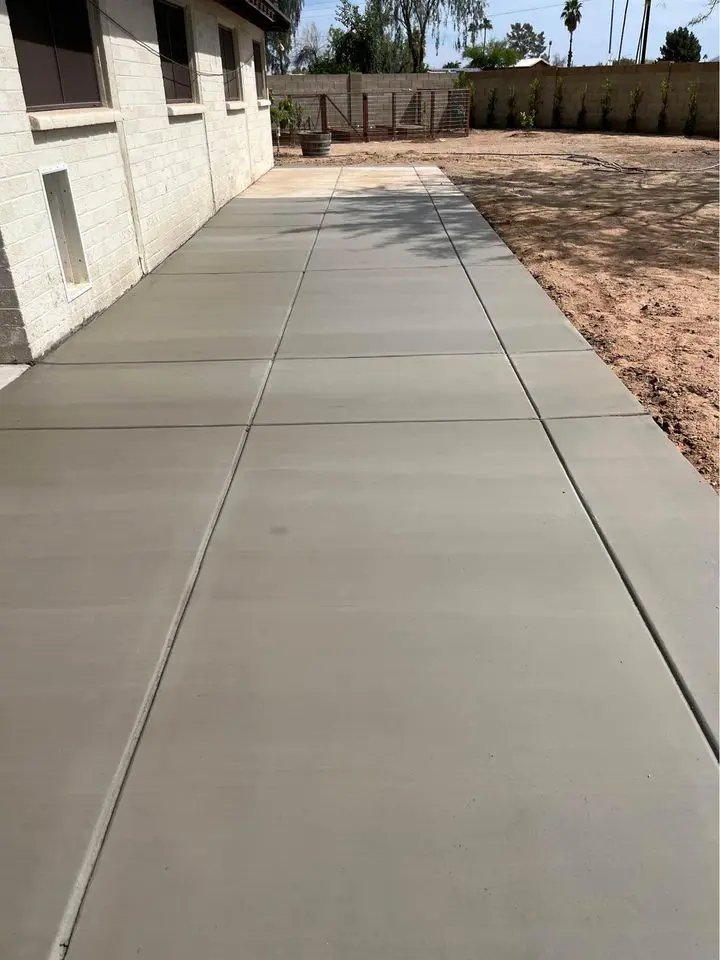 A newly poured concrete walkway, crafted by Bucks Concrete Craft, runs alongside a white brick house in Buckeye, AZ, extending towards a wooden gate. The surrounding area has bare soil and a few scattered trees in the background. The sky is clear.