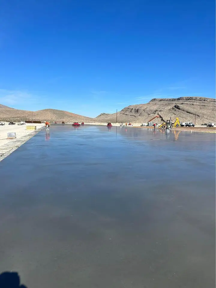 A large, smooth concrete slab stretches toward distant mountains under a clear blue sky in Buckeye, AZ. Workers and construction equipment are seen in the background, with piles of material on the left side—an impressive scene courtesy of Bucks Concrete Craft's expertise as a commercial concrete contractor.