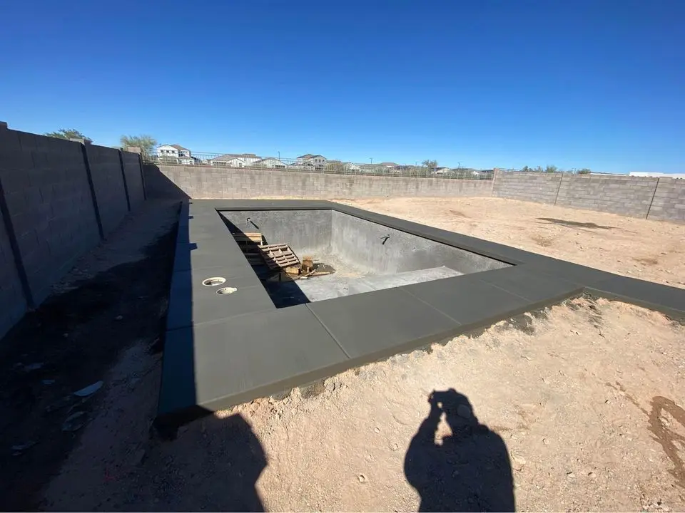 A partially constructed pool with concrete edges awaits pool deck resurfacing, set in a bare, sandy yard. A shadow of a person taking the picture is visible. The background features a high concrete wall and distant houses under a clear blue sky.