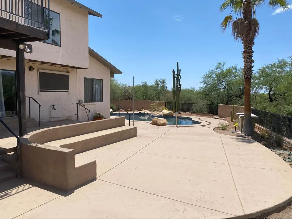 A backyard scene in Buckeye, Arizona, features a swimming pool with a curved design, surrounded by paved decking crafted by a skilled pool deck contractor. Desert plants, including a tall cactus and palm trees, complement the view of the house with steps and balcony under a clear blue sky.