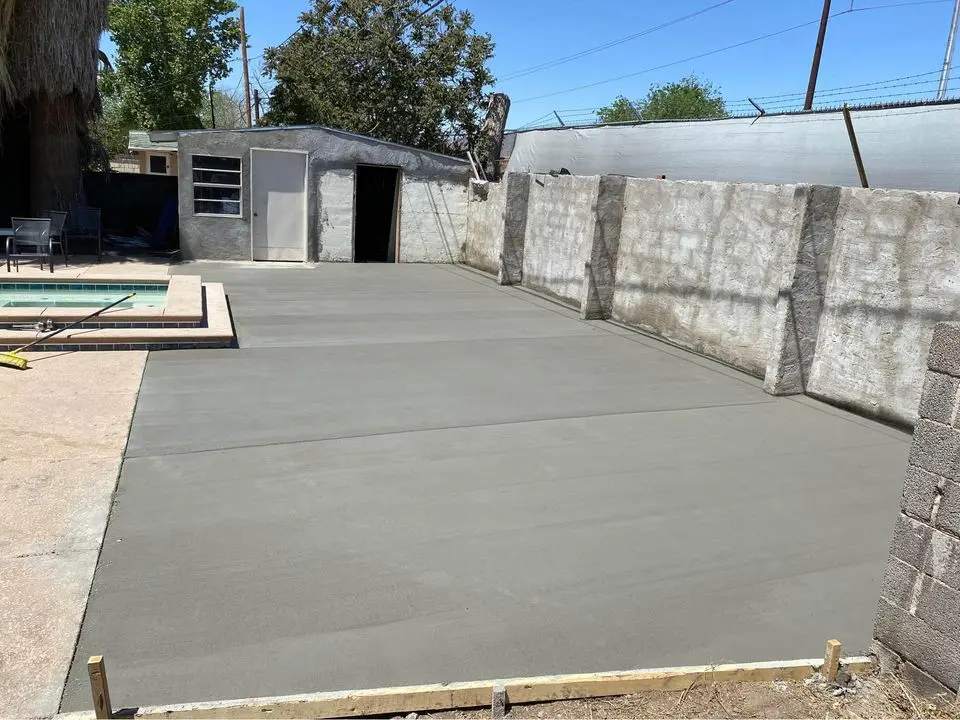 A newly laid concrete patio next to an empty swimming pool hints at a recent pool deck repair. Surrounding walls and wooden forms outline the edges, while in the background, a small building and tree stand under a clear blue sky.
