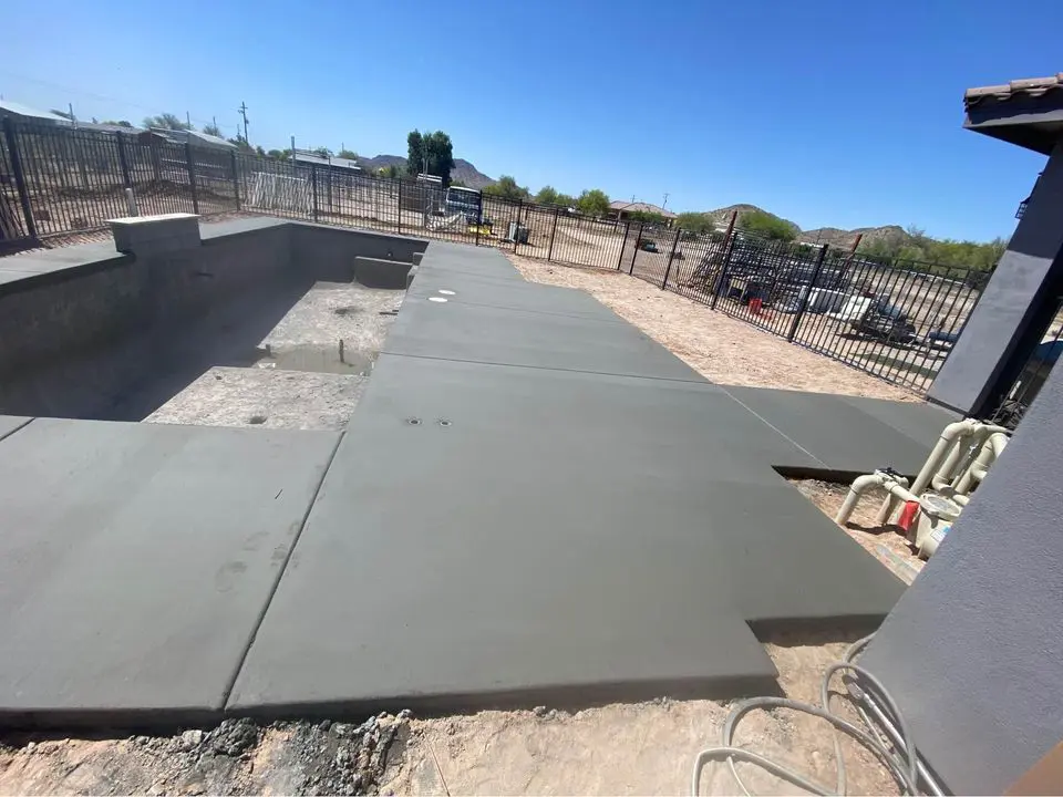 Concrete patio in a backyard under construction in Litchfield Park, featuring a large, partially built pool. The surroundings include fences, desert landscaping, and scattered construction materials. Enjoy the clear blue sky overhead while considering a free estimate from our concrete contractor.