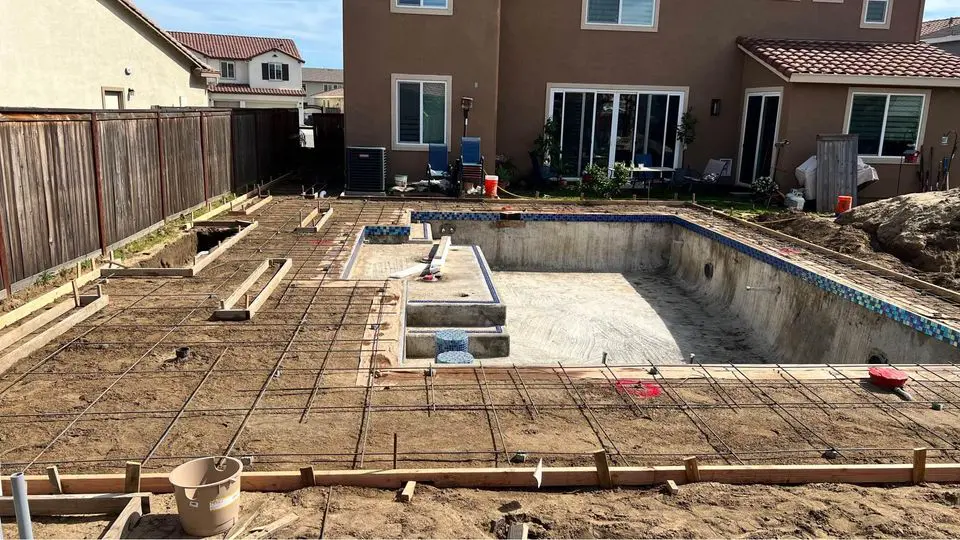 The construction site for an in-ground backyard swimming pool showcases the meticulous work of Goodyear AZ's top concrete contractors, with visible concrete walls and rebar. The yard is framed by wooden fences, against a backdrop of two-story houses under a clear sky.