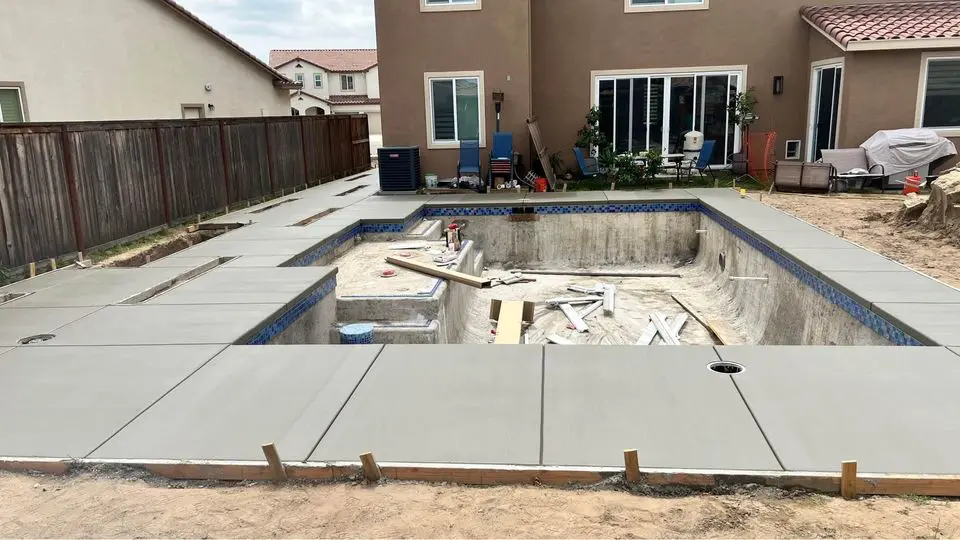 A rectangular backyard pool under construction, surrounded by fresh concrete paving. The pool is empty, with tools and materials inside, waiting for the pool deck contractor to finish. The brown house in the background features large sliding glass doors and several windows.
