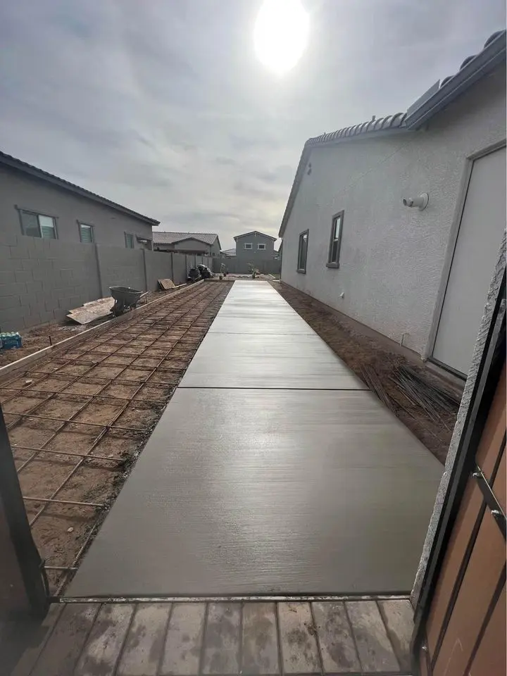 A freshly poured concrete pathway by Bucks Concrete Craft stretches between two houses in Buckeye, AZ, under a cloudy sky with the sun partially visible. Dirt and construction materials lay on either side, showcasing the precision of skilled concrete contractors.