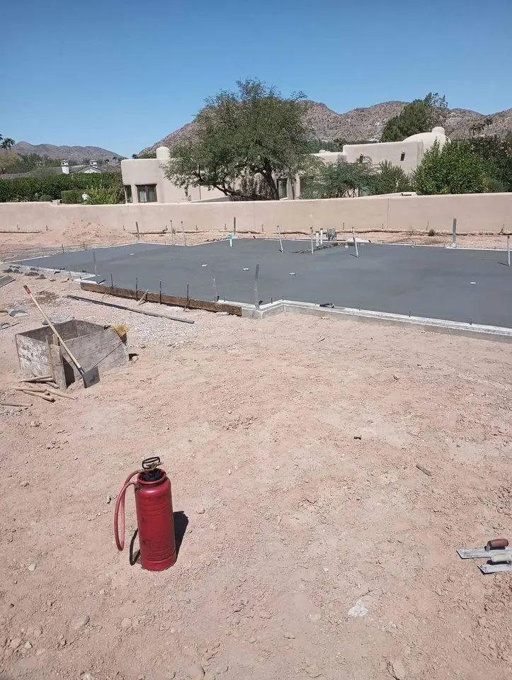 A construction site in Buckeye AZ boasts a newly poured concrete foundation in a sandy area. In the foreground, a red fire extinguisher stands ready. A tree and building can be seen, with mountains rising in the background under a clear blue sky.