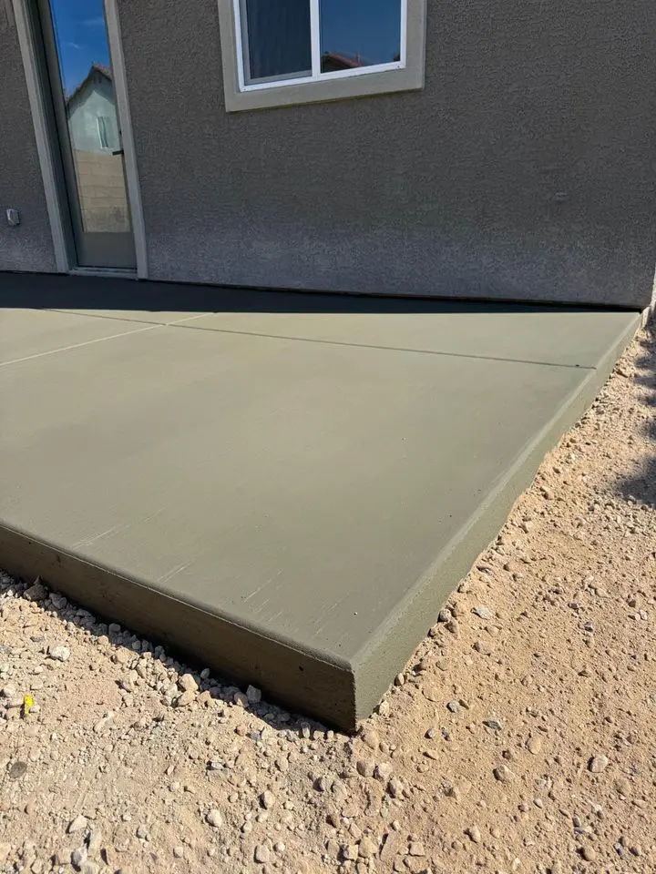 A freshly poured concrete patio slab in front of a door, adjacent to a gray house wall, showcases the skill of local concrete contractors. The ground around the slab consists of loose gravel, while the patio boasts visible sharp edges and a smooth surface, reflective of Buckeye AZ's meticulous craftsmanship.