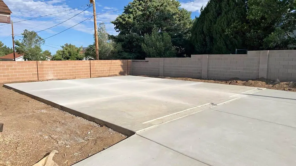 A freshly poured concrete pad in a Buckeye AZ backyard, surrounded by dirt and enclosed by a brick wall. Trees and power lines are visible in the background under a partially cloudy sky, showcasing the quality work of foundation contractors.