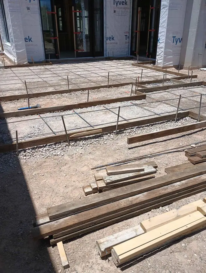 A construction site in Buckeye, AZ showcases the foundation layout with metal rebar grids and wooden planks ready for concrete pouring. Skilled concrete contractors work efficiently as partially constructed walls are covered with Tyvek sheeting.