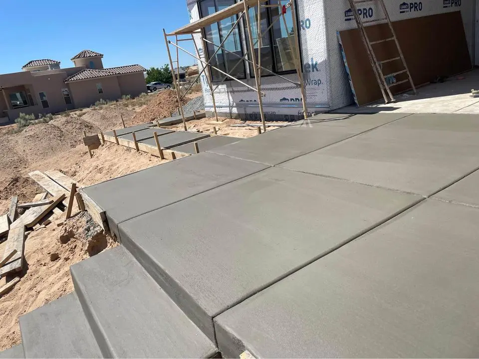 Freshly poured concrete steps and pathway lead to the entrance of a house under construction in Buckeye, AZ, with scaffolding and building materials visible. Concrete contractors work diligently as a modern house stands against the clear blue sky in the background.