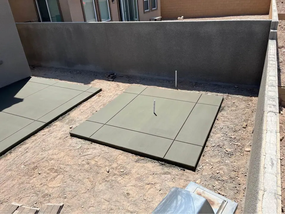 A backyard in Litchfield Park boasts newly laid rectangular patio slabs on bare earth, all part of a concrete construction project. Surrounded by sturdy concrete walls, a metal pipe emerges from the center, with residential buildings providing a picturesque backdrop.