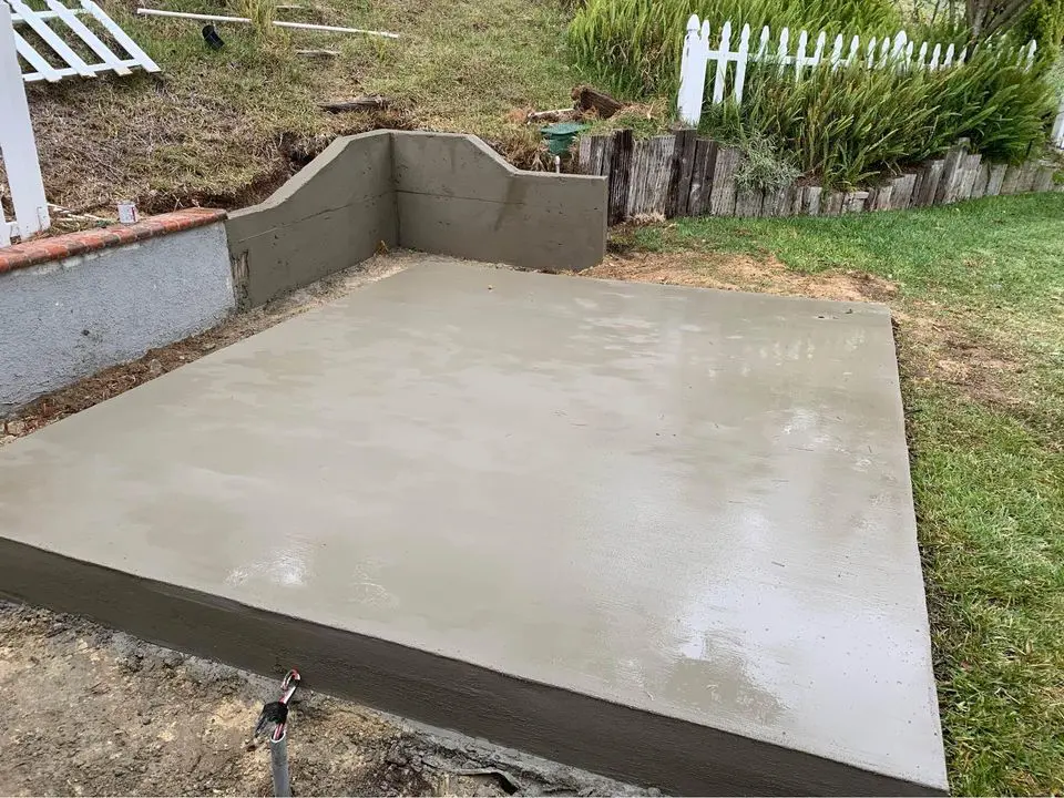 A freshly poured concrete slab lies adjacent to a small grassy area and a white picket fence, the craftsmanship evident—a testament to skilled concrete contractors in Goodyear, AZ. In the background, a raised section of concrete and scattered garden tools complete the scene.