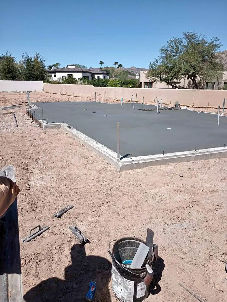 A freshly poured concrete foundation with smooth edges sits on a construction site in Buckeye, AZ. Surrounding the foundation are scattered tools and equipment from local concrete contractors. The area is bordered by sand and a few trees, with houses visible in the background under a clear blue sky.