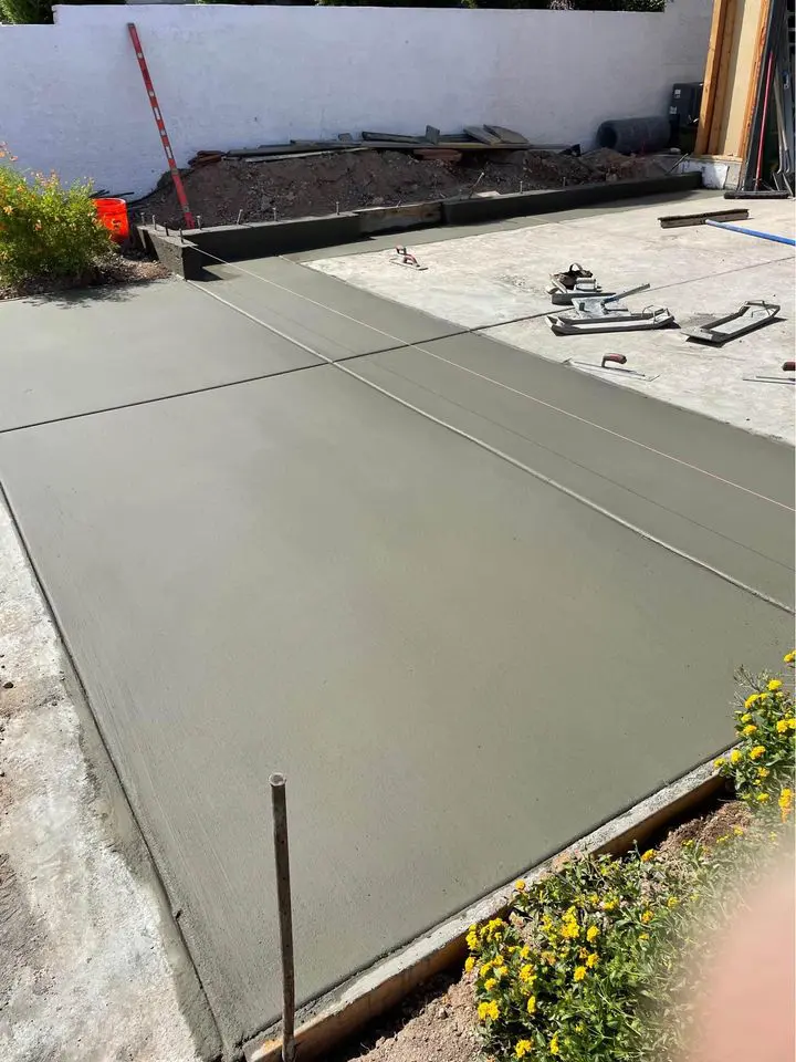 A freshly poured concrete slab, crafted by skilled Concrete Patio Contractors in West Valley, boasts a smooth surface sectioned by control joints. The surrounding area has construction tools, small yellow flowers, and an unfinished boundary with soil and equipment in the background.