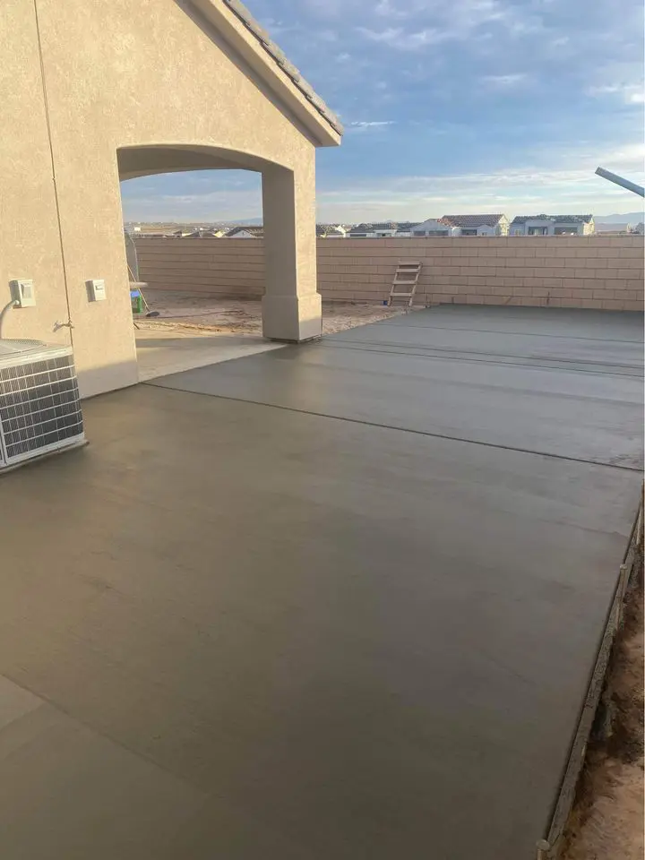 A newly poured concrete patio graces a house in Litchfield Park with an arched entrance. The sky is clear, and a low brick wall embraces the area. A ladder leans against the wall near a visible air conditioning unit, crafted to perfection by a skilled concrete contractor offering free estimates.