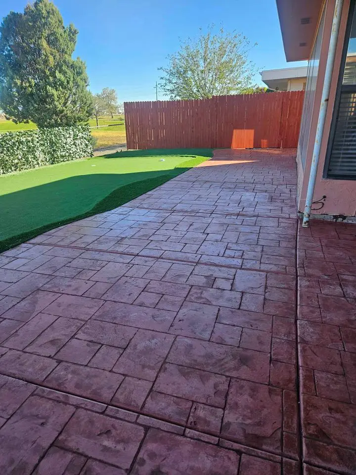 The backyard in West Valley features a red brick-patterned patio, expertly crafted by Concrete Patio Contractors, complemented by artificial green turf and a wooden privacy fence. A tree stands in the background near the house with a window on the right, all under a clear blue sky.