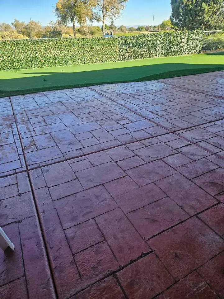 A wet, patterned concrete patio crafted by skilled concrete contractors in Buckeye AZ, with a green artificial turf area in the background. A hedge effortlessly separates the space from the driveways, trees, and sky in the distance.