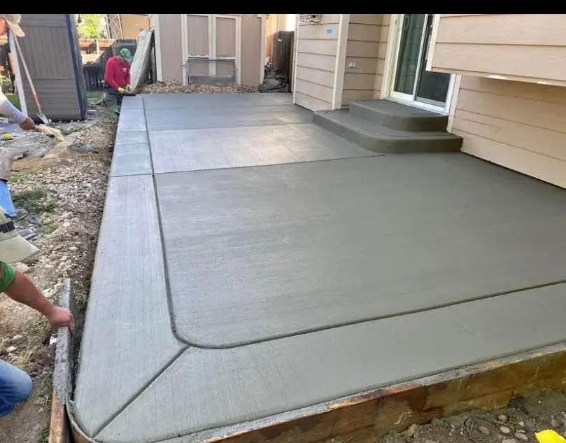A freshly poured concrete patio with smooth edges graces a Litchfield Park backyard. Workers from a skilled concrete contractor are smoothing the surface. Adjacent steps lead to the house entrance, and a small pile of debris and tools is visible in the background.