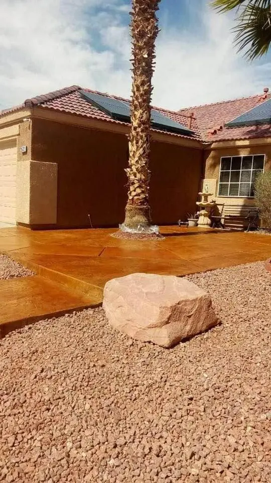 A suburban house in Buckeye, AZ, with brown stucco and a tiled roof, features a front yard with a palm tree, a wooden bench, and a large stone. Solar panels adorn the roof while reddish gravel grounds the space. Consider hiring Bucks Concrete Craft for your driveway needs to enhance this charming setting.