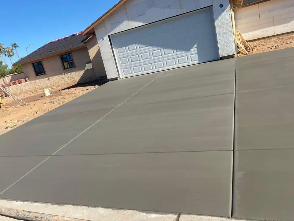 A freshly poured concrete driveway leads to a two-car garage of a single-story house under construction, showcasing the expert work of concrete contractors. The house has unfinished walls, and the surrounding yard is bare soil scattered with some building materials.