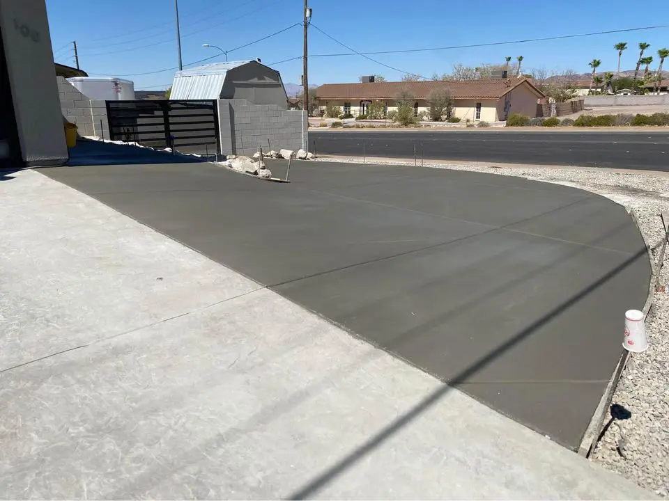 A newly paved concrete driveway, crafted by a skilled Buckeye AZ contractor, curves towards the road. Set in a residential area with a fence and houses under a clear blue sky, small rocks are scattered alongside this testament to quality concrete construction.