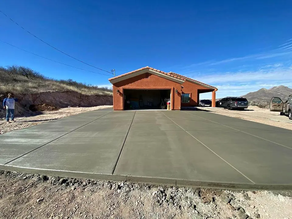 A newly paved concrete driveway by a skilled contractor leads to a red brick house with an open garage. Two cars are parked on the side, and a person stands nearby under the clear blue sky with hills in the background, capturing a serene day in Buckeye, AZ.