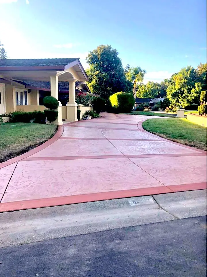 A wide, pinkish driveway crafted by a skilled concrete contractor leads to a house with a tiled roof, surrounded by a well-maintained lawn and trees. The sky is clear, and the sun casts long shadows across the property in Litchfield Park.