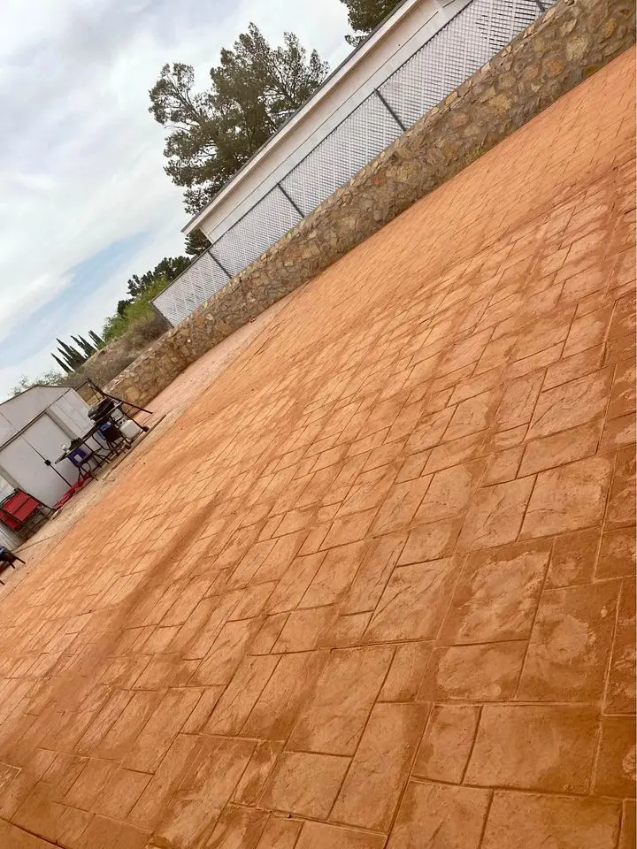 A large, paved courtyard features reddish-brown tiles against a stone wall. To the left, a few outdoor tables and chairs offer a cozy spot for relaxation. Nestled under a cloudy Buckeye, AZ sky, trees provide a natural backdrop to this charming space crafted by an expert concrete construction team.
